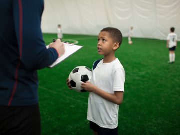 Adorable football player with ball talking to his trainer on pitch after game